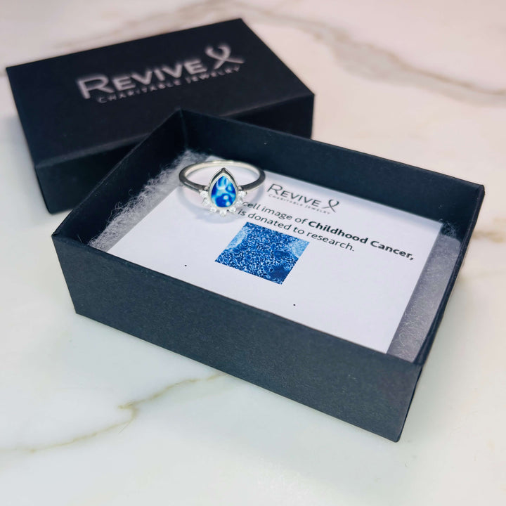Sterling silver teardrop shaped ring with half halo of cubic zirconia stones and childhood cancer cell image under resin pendant with product packaging