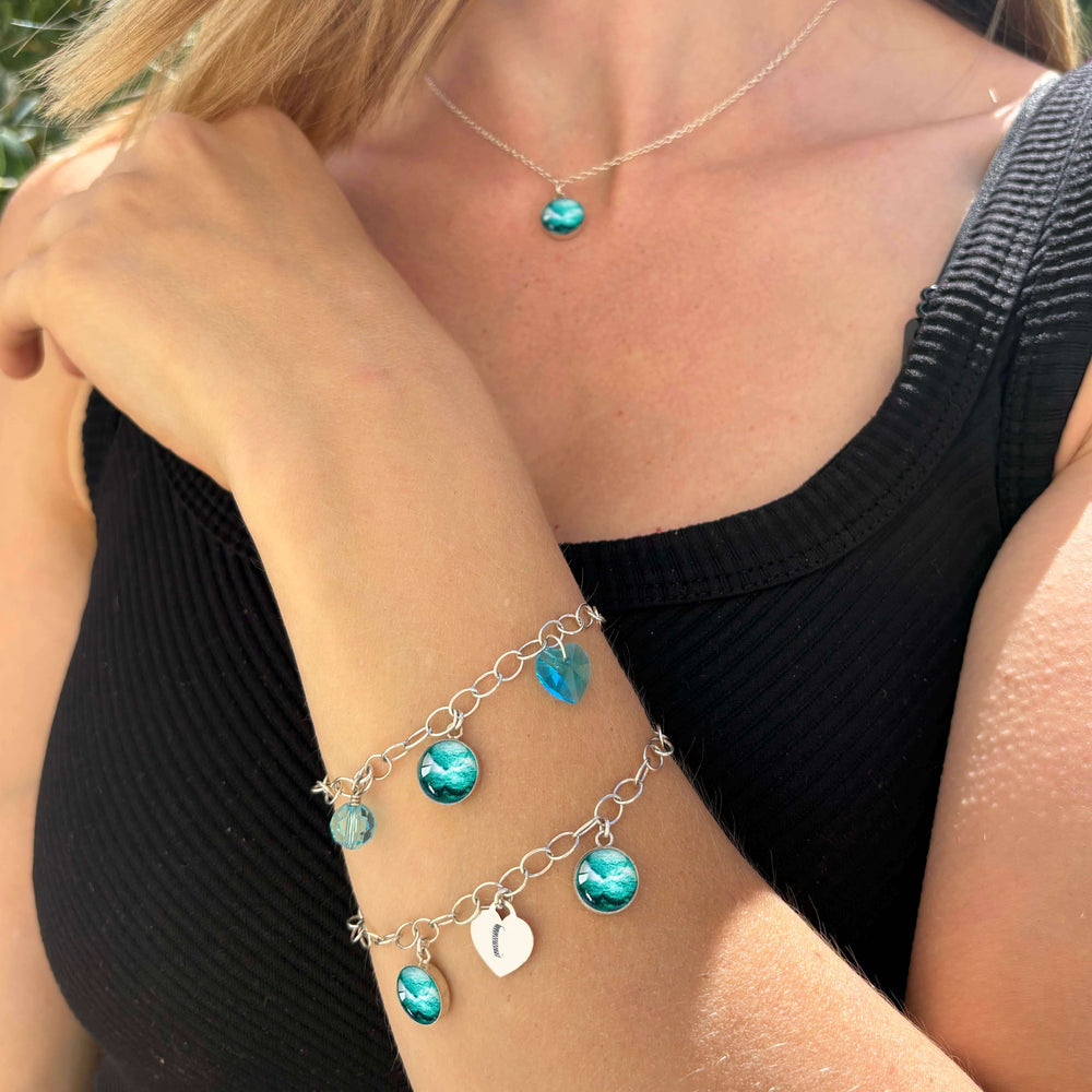 Model wearing silver bracelets with teal embryo images, blue crystals, and a heart-shaped charm engraved with "C"