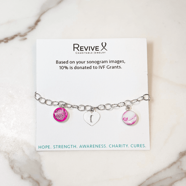 Silver bracelet with pink pendants showing the IVF journey from embryo to baby and an engraved heart-shaped charm on a marble background