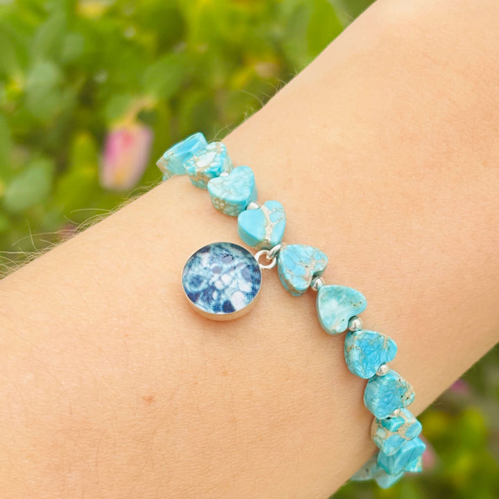 close up of model wearing blue sea sediment jasper heart shaped bead bracelet with silver accent beads and round ovarian cancer cell image in resin pendant